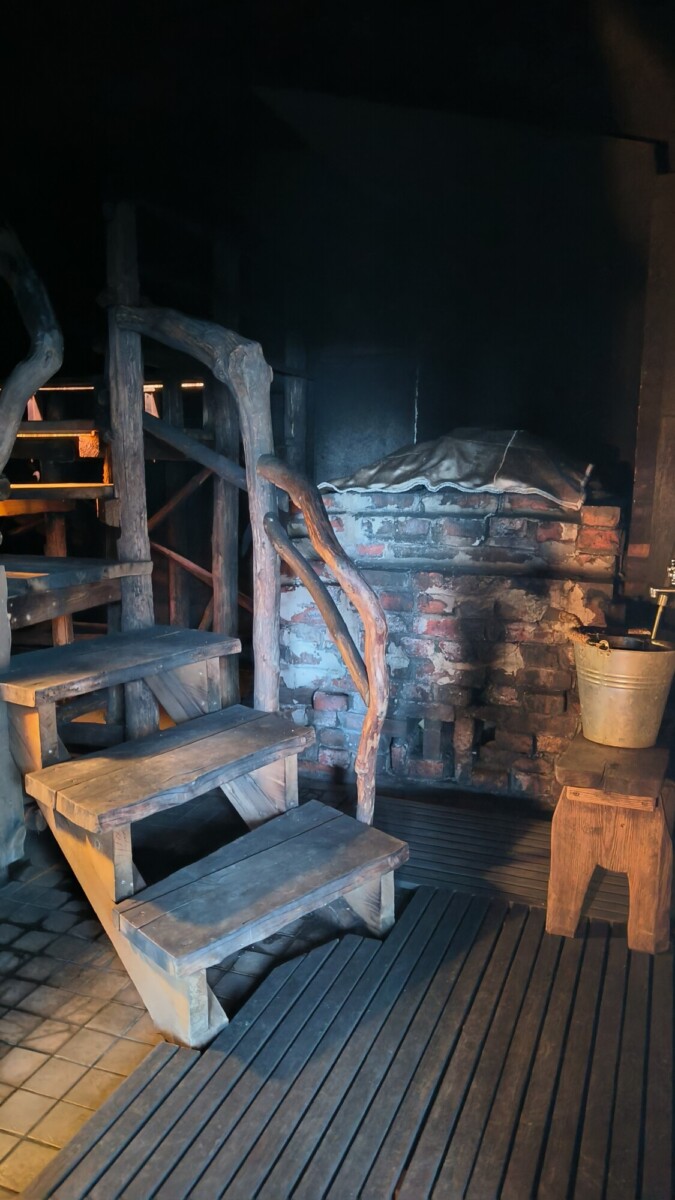 Pienen tiilitakan vieressä on maalaismainen puuportaikko, jossa on karkeasti veistetyt kaiteet. Siellä on puinen jakkara, jonka päällä on metalliämpäri. Lämmin, hämärä valaistus luo kodikkaan, vanhanaikaisen tunnelman.
