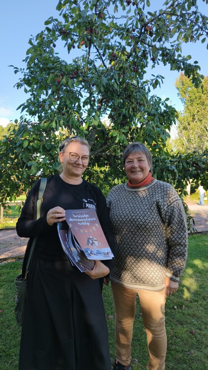 Kaksi naista seisoo ulkona lehtimäisen puun edessä. Toisella on pino kuvitettuja vihkosia, ja hänellä on silmälasit ja musta toppi; toisella on kuvioitu villapaita. On aurinkoinen päivä, ruoho on vihreää ja taivas sininen.