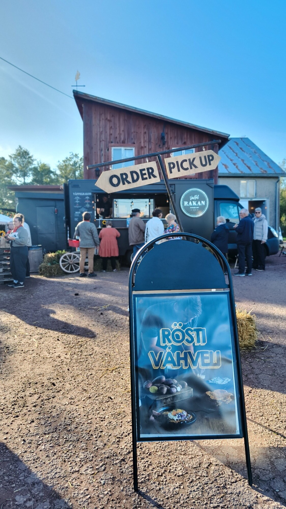 Joukko ihmisiä jonottaa ulkona sijaitsevan ruokakojun ääressä maalaismaisen puurakennuksen edessä. Etualalla on kyltti, jossa on kuvia ruoasta ja jossa lukee RÖSTI VÄHVELI. Kojun yläpuolella on kyltit ORDER ja PICK UP.