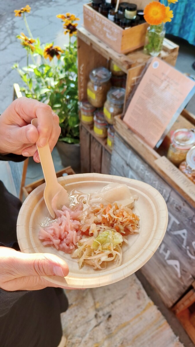 Henkilö pitelee lautasta puuhaarukalla ja maistelee marinoituja vihanneksia ulkona sijaitsevalla myyntikojulla, jonka taustalla näkyy purkkeja, kukkia ja ruokalista.