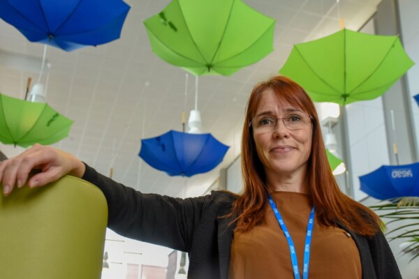 Punatukkainen, silmälasipäinen nainen hymyilee, hänellä on ruskea toppi ja sininen nauha, ja hän seisoo sisätiloissa, kun taustalla on katosta ylösalaisin roikkuvia vihreitä ja sinisiä sateenvarjoja.