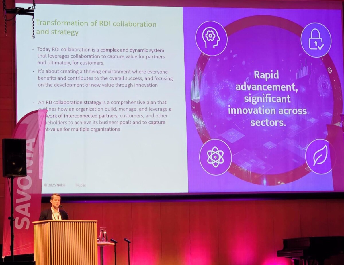 Puhuja seisoo puhujakorokkeella suuren näytön edessä, jossa on esitys TKI-yhteistyöstä ja -strategiasta, ja siinä on violettia grafiikkaa ja tekstiä nopeasta edistyksestä ja innovoinnista. Vasemmalla näkyy SAVONIA-banneri.