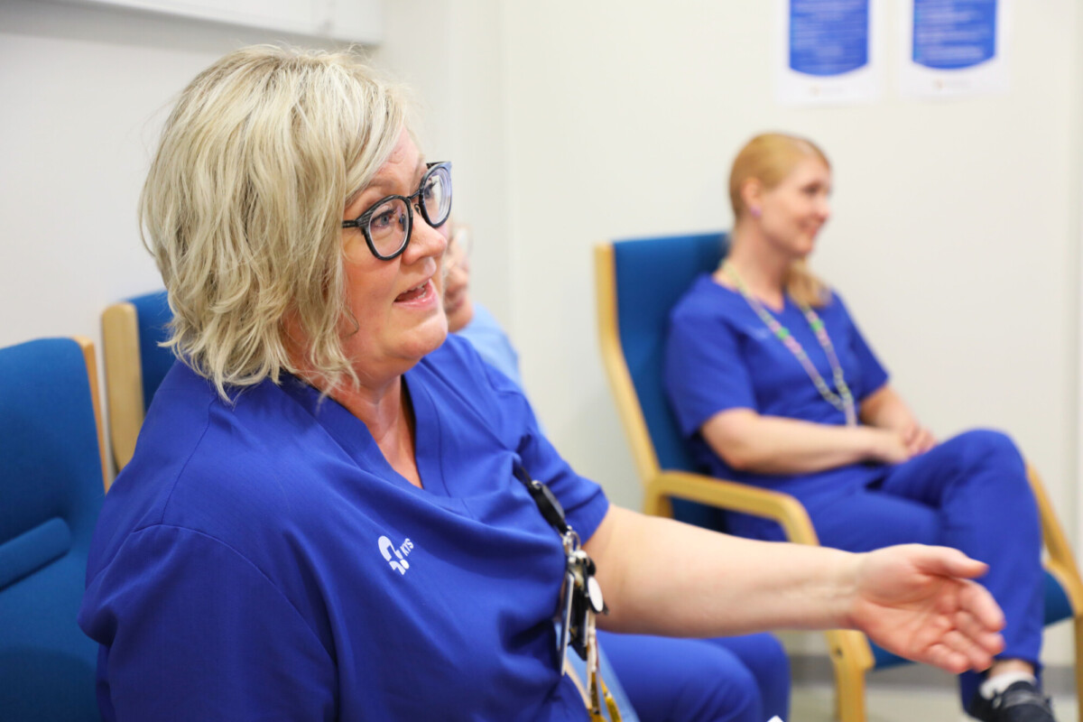 Siniseen hoitohaalariin pukeutunut nainen tekee eleitä puhuessaan ja istuu tuolilla. Taustalla, hieman epätarkasti, istuu toinen henkilö samanlaisissa leikkaushousuissa kirkkaasti valaistussa huoneessa.