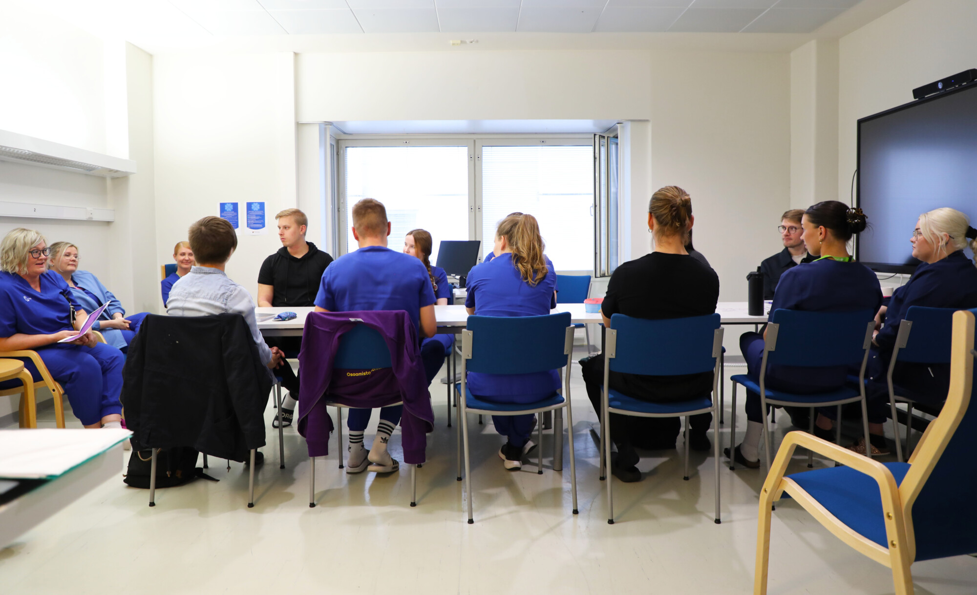 Joukko ihmisiä, joista osa on pukeutunut sinisiin ja osa mustiin työasuihin, istuu pöydän ympärillä kirkkaasti valaistussa kokoushuoneessa keskustelemassa. Oikealla olevalla seinällä roikkuu suuri monitori.