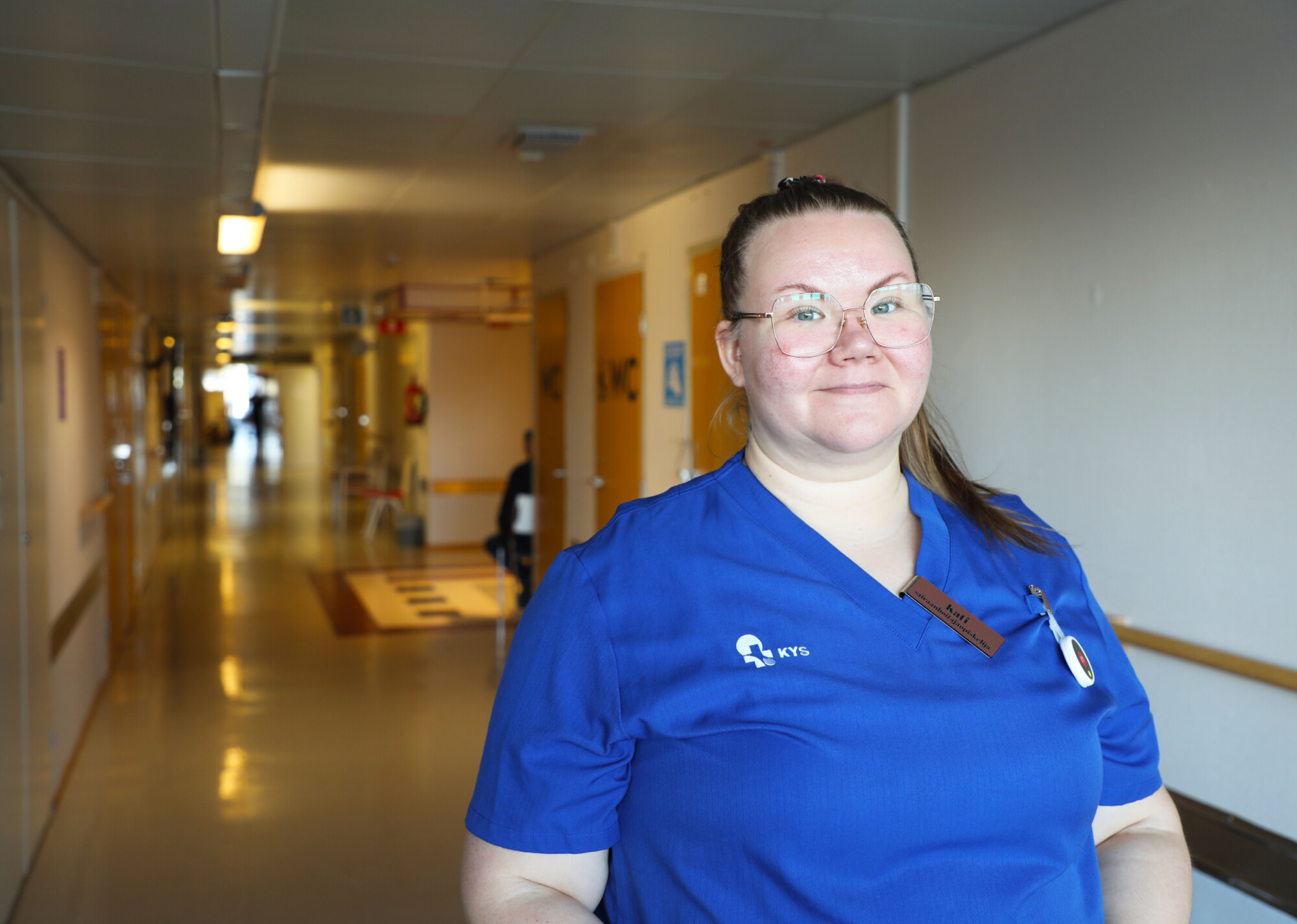 Sairaanhoitaja sinisessä asussa seisoo kirkkaasti valaistulla sairaalan käytävällä ja hymyilee kameralle. Käytävä ulottuu taustalle, ja seinillä näkyy ovia ja lääketieteellisiä kylttejä.