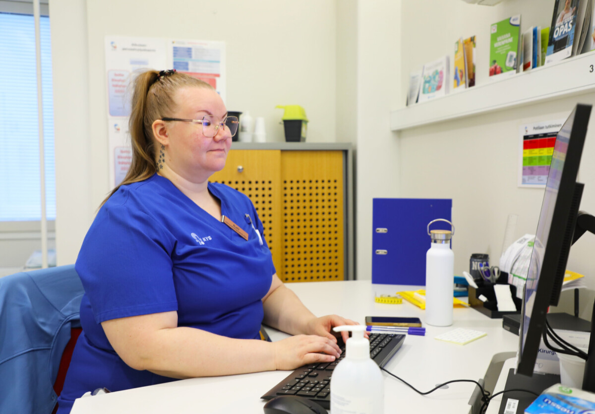 Sinisiin leikkaushameisiin pukeutunut terveydenhuollon työntekijä istuu työpöydän ääressä ja kirjoittaa tietokoneella. Työpöydällä on toimistotarvikkeita, vesipullo ja käsihuuhde. Terveysalan esitteitä ja asiakirjoja on näkyvissä hyllyissä ja seinillä.