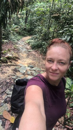 Nainen ottaa reppu selässään selfien kivisellä, kapealla polulla, jota ympäröi tiheä, vihreä viidakon lehdistö. Naisella on vaalea iho ja taaksepäin sidotut punertavat hiukset, ja hänellä on yllään violetti paita.