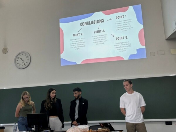 Neljä henkilöä seisoo luokkahuoneen edessä pitämässä esitelmää. Heidän takanaan olevalle seinälle heijastetaan johtopäätösdia, jossa on neljä pistettä. Kello näyttää, että kello on noin 1:15.