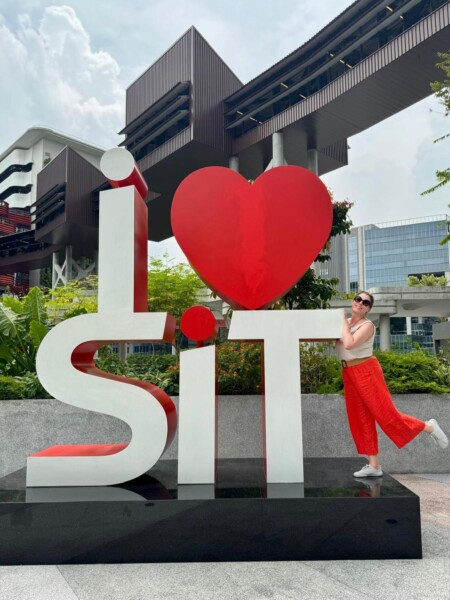 Punaisiin housuihin ja aurinkolaseihin pukeutunut henkilö poseeraa ulkona suuren "I ♥ SiT" -kyltin vieressä, taustalla on moderneja rakennuksia ja vehreyttä.