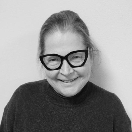 Nainen, jolla on pitkät taakse vedetyt hiukset, suuret mustat silmälasit ja tumma pusero, hymyilee kameraan päin vaaleaa taustaa vasten.