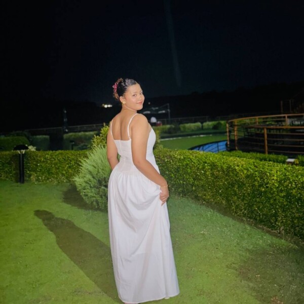 Valkoiseen hihaton mekkoon pukeutunut nainen seisoo ulkona nurmikolla yöllä, hieman poispäin, mutta katsoo takaisin kameraan. Taustalla on siististi leikattuja pensaita ja uima-allas tumman taivaan alla.