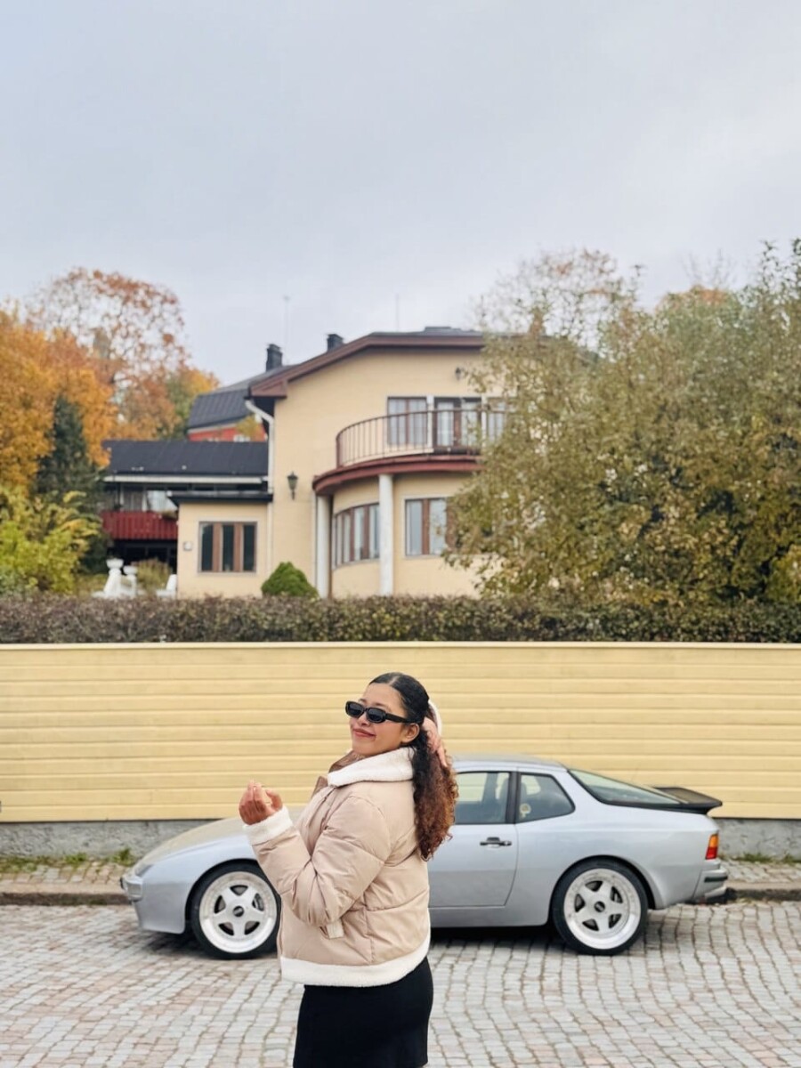 Aurinkolaseihin ja kevyeen takkiin pukeutunut nainen poseeraa ja hymyilee hopeisen urheiluauton edessä, joka on pysäköity beigen aidan viereen, ja taustalla on suuri talo ja syksyisiä puita.