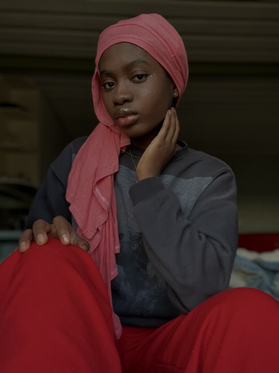 Vaaleanpunaiseen huiviin ja punaisiin housuihin pukeutunut nuori nainen istuu sisätiloissa ja painaa kätensä kasvoilleen. Hän katsoo suoraan kameraan rauhallinen, luottavainen ilme. Tausta on pehmeästi sumea.