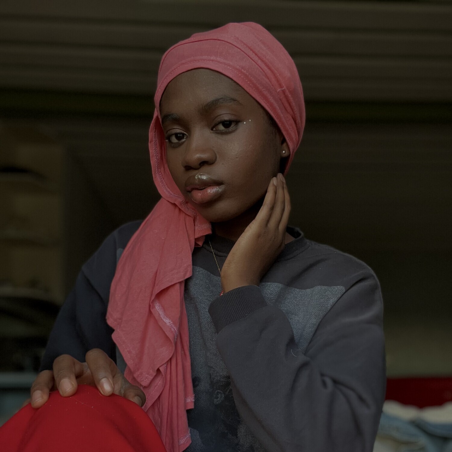 Vaaleanpunaiseen huiviin ja punaisiin housuihin pukeutunut nuori nainen istuu sisätiloissa ja painaa kätensä kasvoilleen. Hän katsoo suoraan kameraan rauhallinen, luottavainen ilme. Tausta on pehmeästi sumea.