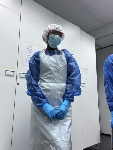 Henkilö, joka on pukeutunut täydellisiin henkilökohtaisiin suojavarusteisiin, kuten hiussuojukseen, kasvonaamariin, suojalaseihin, kaapuun, muoviseen esiliinaan ja sinisiin käsineisiin, seisoo kädet yhteenliitettynä kliinisessä tilassa.