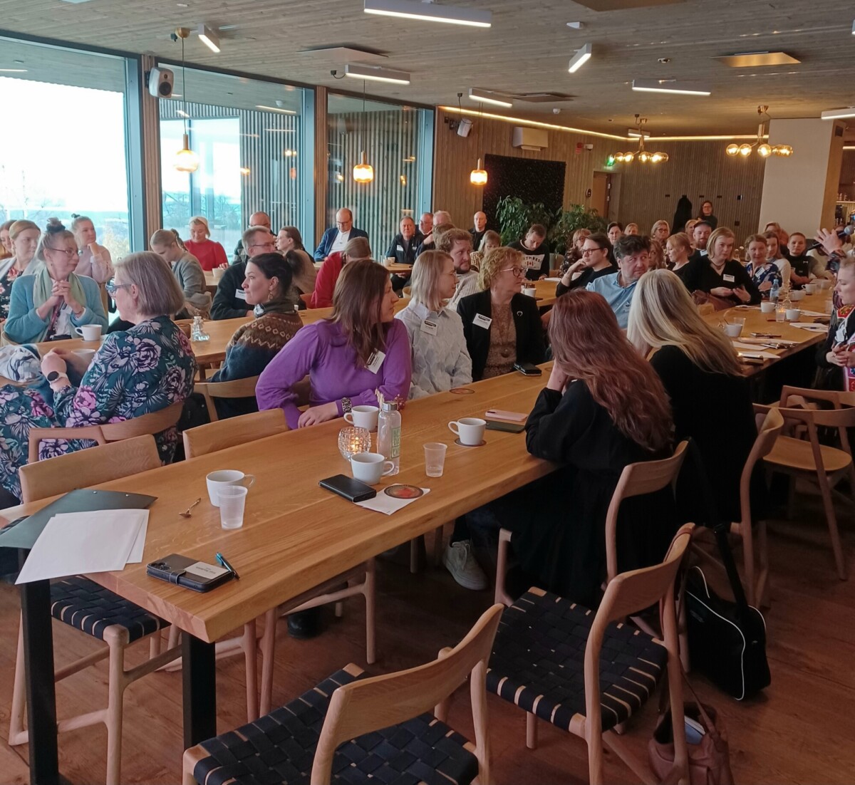 Suuri joukko ihmisiä istuu puisten pöytien ääressä modernissa, hyvin valaistussa huoneessa ja kuuntelee tarkkaavaisesti puhujaa, joka ei ole näkyvissä. Jotkut tekevät muistiinpanoja tai pitävät juomia kädessään. Tunnelma on rento ja sitoutunut.