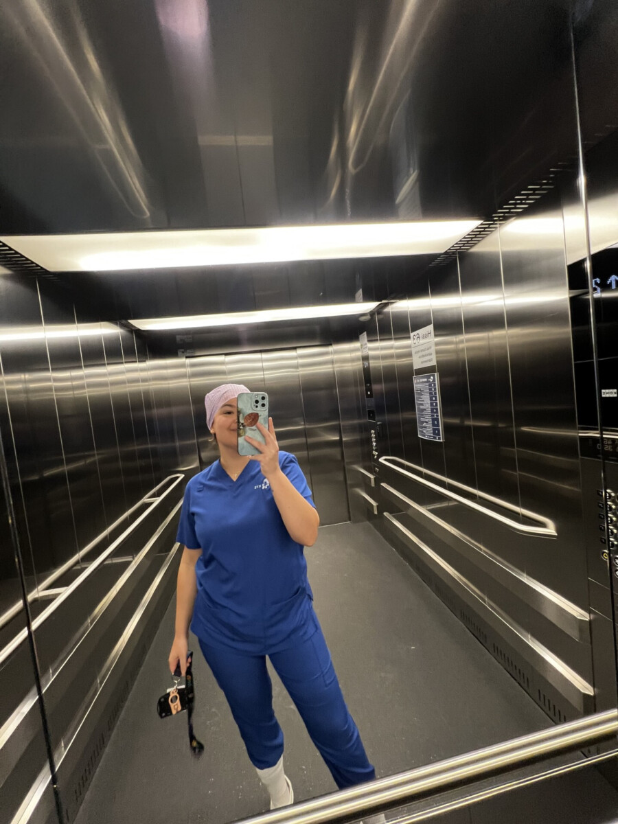 Henkilö, jolla on sininen lääkärinhoitohaalari ja vaaleanvioletti päähine, ottaa peiliselfien ruostumattomasta teräksestä valmistetun hissin sisällä pitäen puhelinta toisessa kädessä ja kameraa toisessa.