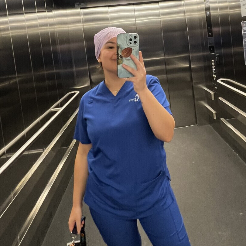 Henkilö, jolla on sininen lääkärinhoitohaalari ja vaaleanvioletti päähine, ottaa peiliselfien ruostumattomasta teräksestä valmistetun hissin sisällä pitäen puhelinta toisessa kädessä ja kameraa toisessa.