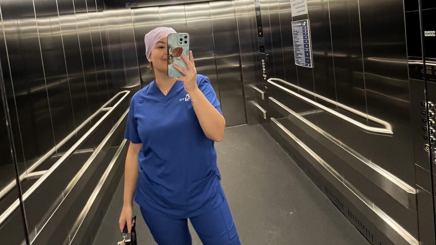 Henkilö, jolla on sininen lääkärinhoitohaalari ja vaaleanvioletti päähine, ottaa peiliselfien ruostumattomasta teräksestä valmistetun hissin sisällä pitäen puhelinta toisessa kädessä ja kameraa toisessa.