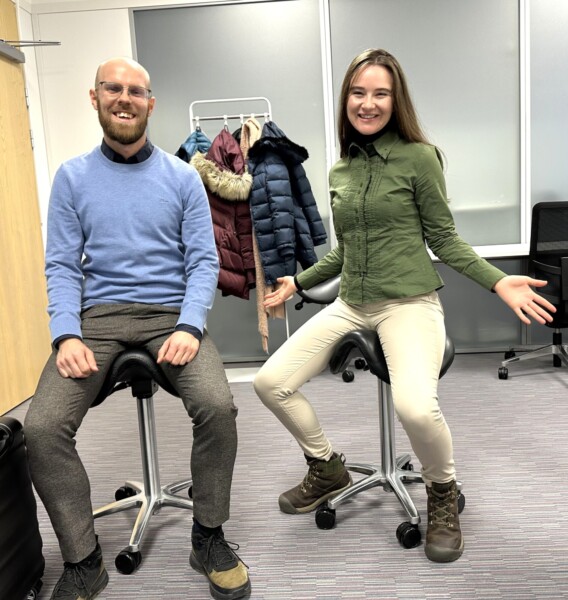 Mies ja nainen hymyilevät ja istuvat satulamallisilla toimistotuolilla modernissa toimistohuoneessa. Takit roikkuvat telineessä heidän takanaan, ja nainen elehtii innostuneesti avosylin.