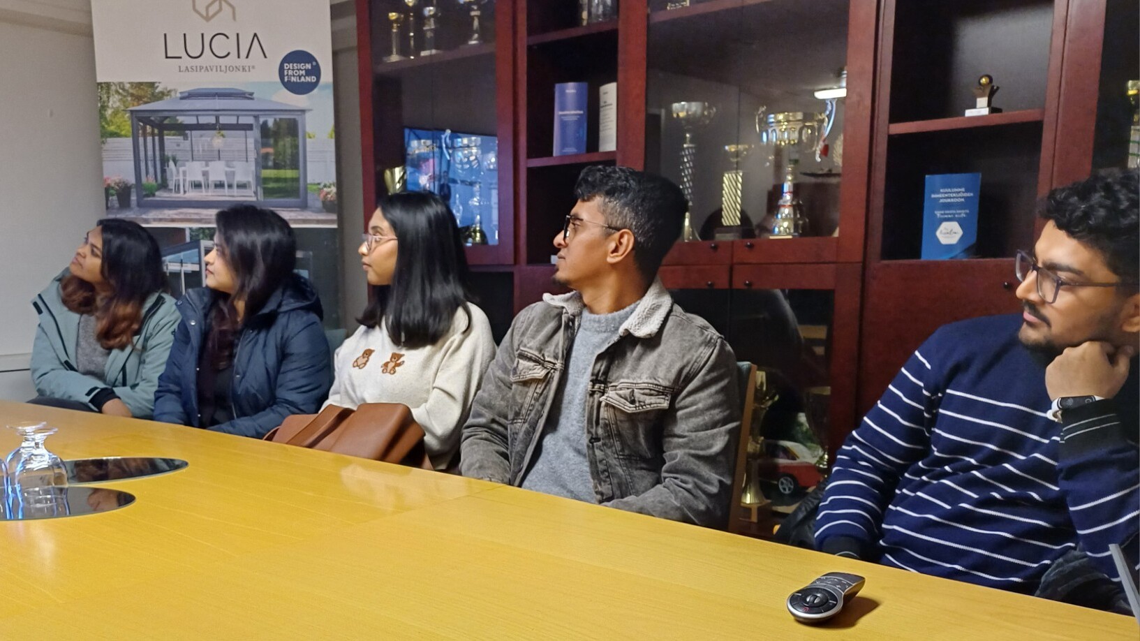 Viisi ihmistä istuu kokouspöydän ääressä ja katselee tarkkaavaisesti esitystä näytöltä, pöydällä on lasitavaraa ja heidän takanaan kaapeissa on pokaaleja.