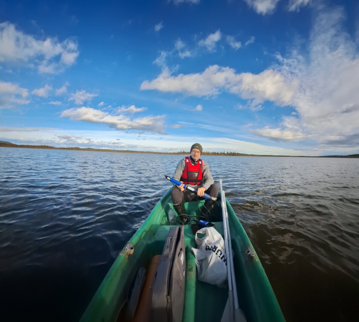 Henkilö, jolla on punainen pelastusliivi ja pipo, istuu vihreässä kanootissa rauhallisella järvellä ja pitää melaa kädessään. Taivas on sininen, ja pilviä on hajallaan, ja kanootissa näkyy pusseja.