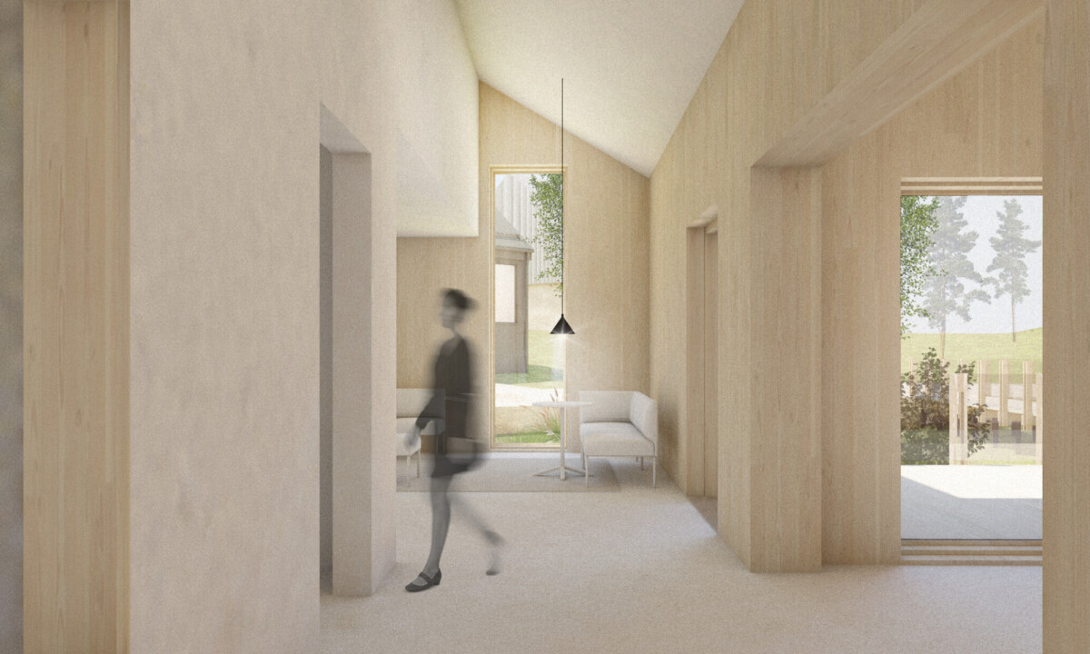 Sumea henkilö kävelee valoisassa, minimalistisessa huoneessa, jossa on vaaleat puuseinät ja korkeat katot. Suuret ikkunat päästävät luonnonvaloa sisään ja paljastavat ulkona olevat vihreät puut ja terassin. Yhden ikkunan vieressä on pieni pöytä ja tuolit.