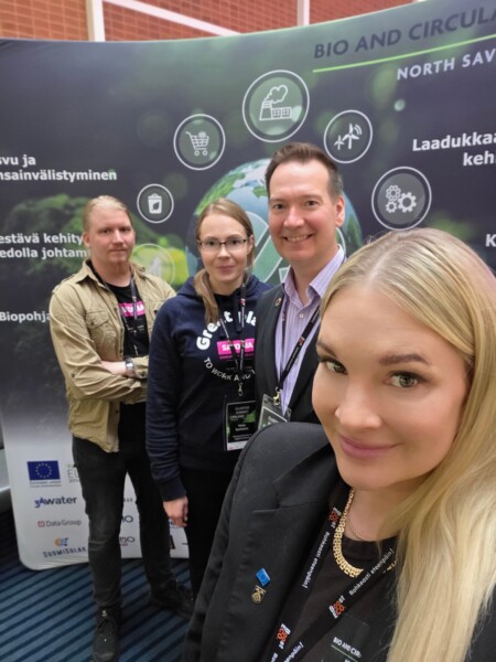 Neljä ihmistä seisoo sisätiloissa konferenssitaustan edessä, jossa on logoja ja tekstiä bio- ja kiertotaloudesta. Kolme hymyilee kameralle, kun taas yksi näyttää vakavalta. Kaikilla on yllään konferenssimerkit ja liikemiesten asu.