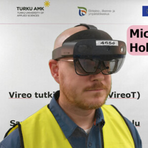 Henkilö, jolla on näkösuojainliivit ja Microsoft Hololens 2 -kuulokkeet, seisoo valkoisen taustan edessä, jossa on Savonian, Turun ammattikorkeakoulun ja Euroopan unionin logoja.
