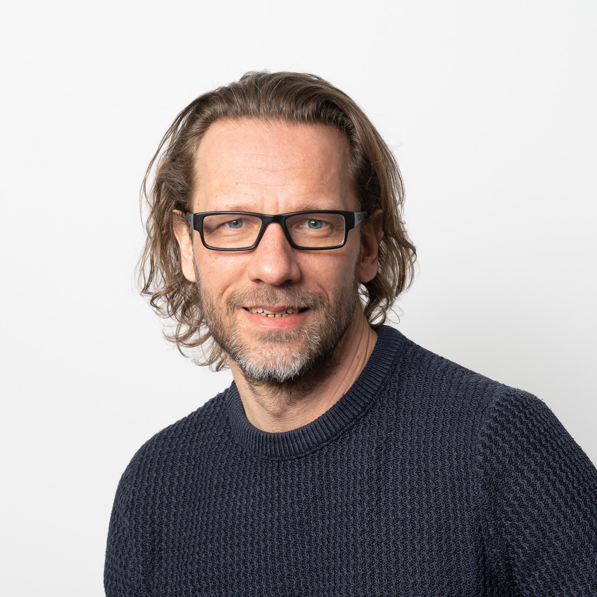 Mies, jolla on olkapäiden pituinen aaltoileva tukka, silmälasit ja parta, on pukeutunut tummaan puseroon ja hymyilee hieman valkoista taustaa vasten.