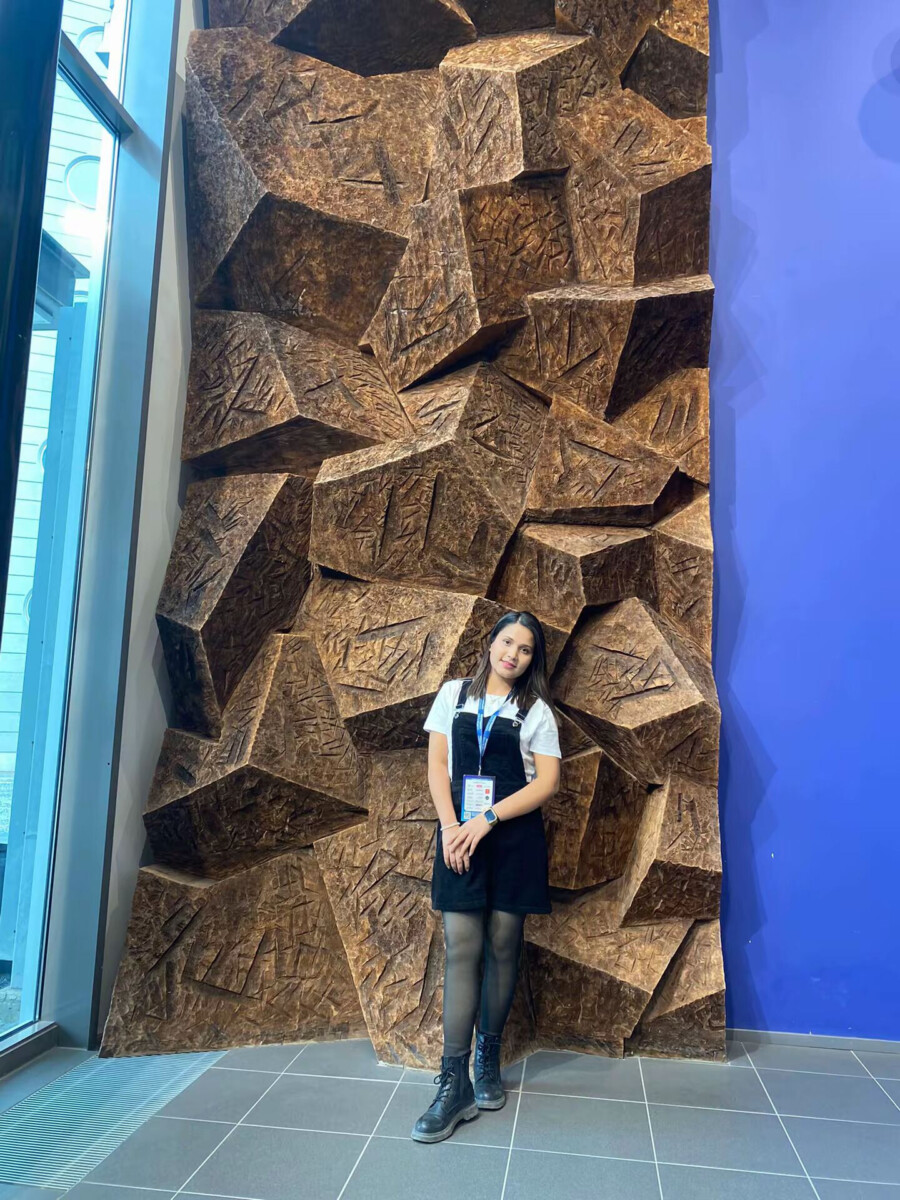 Nainen seisoo ja hymyilee korkean, teksturoidun geometrisen seinäinstallaation edessä, jossa on ruskeita, kulmikkaita muotoja, suuren ikkunan ja sinisen seinän vieressä. Naisella on yllään valkoinen paita, musta mekko ja saappaat.