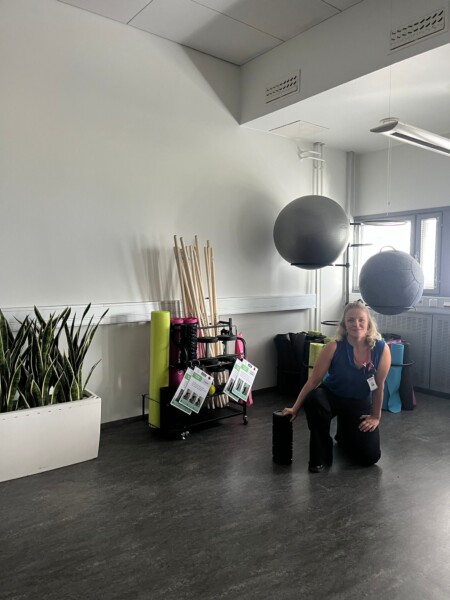 Nainen polvistuu kuntosalin lattialla, kädessään musta vaahtomuovirulla. Hänen takanaan on jumppapalloja, vaahtomuovimattoja, pystyssä olevia puukeppiä ja istutusastia, jossa on korkeita vihreitä kasveja.