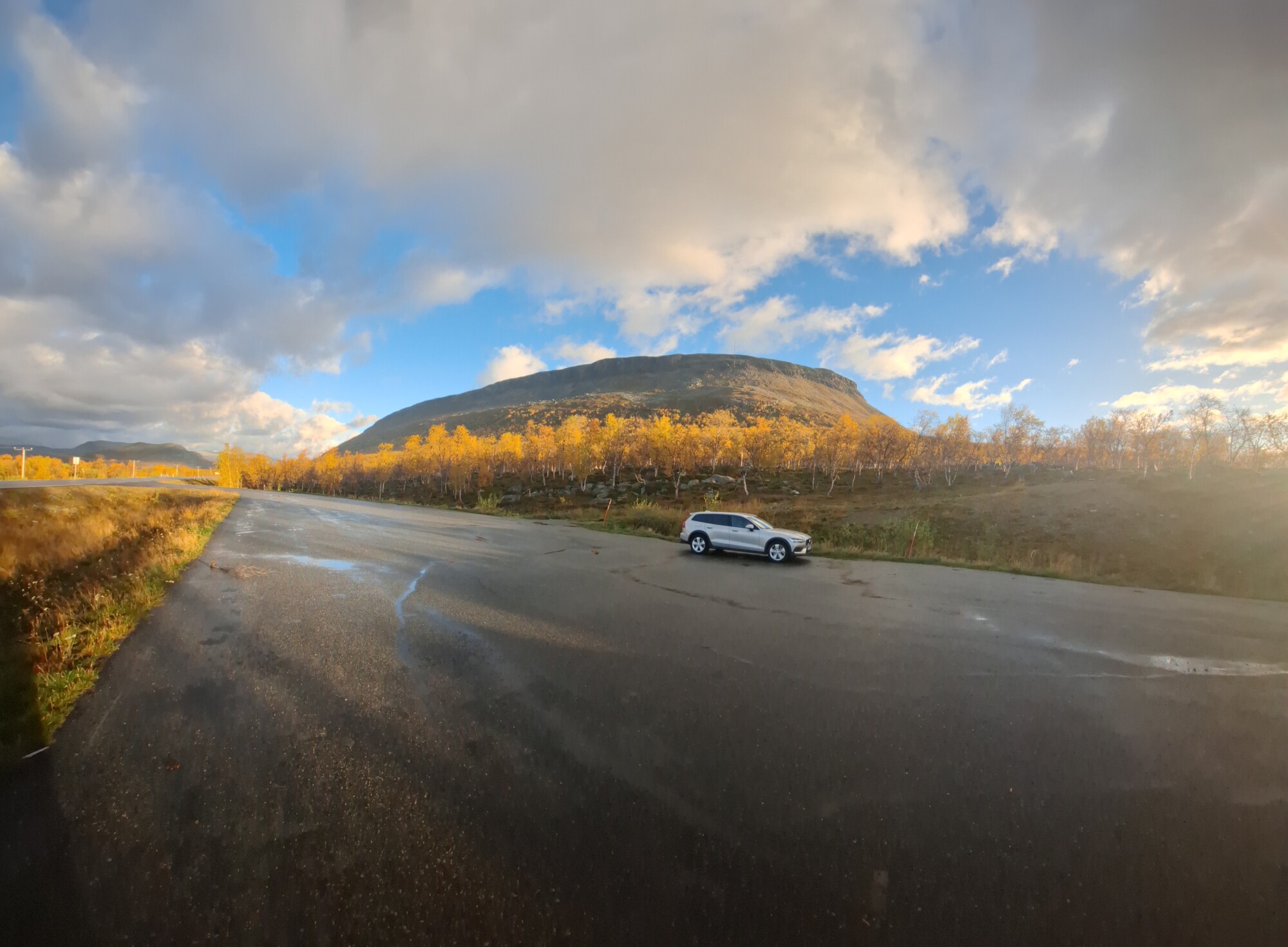 Valkoinen auto on pysäköity märälle, tyhjälle päällystetylle alueelle, jonka taustalla on syksynvärisiä puita ja korkea, kallioinen kukkula osittain pilvisen taivaan alla.