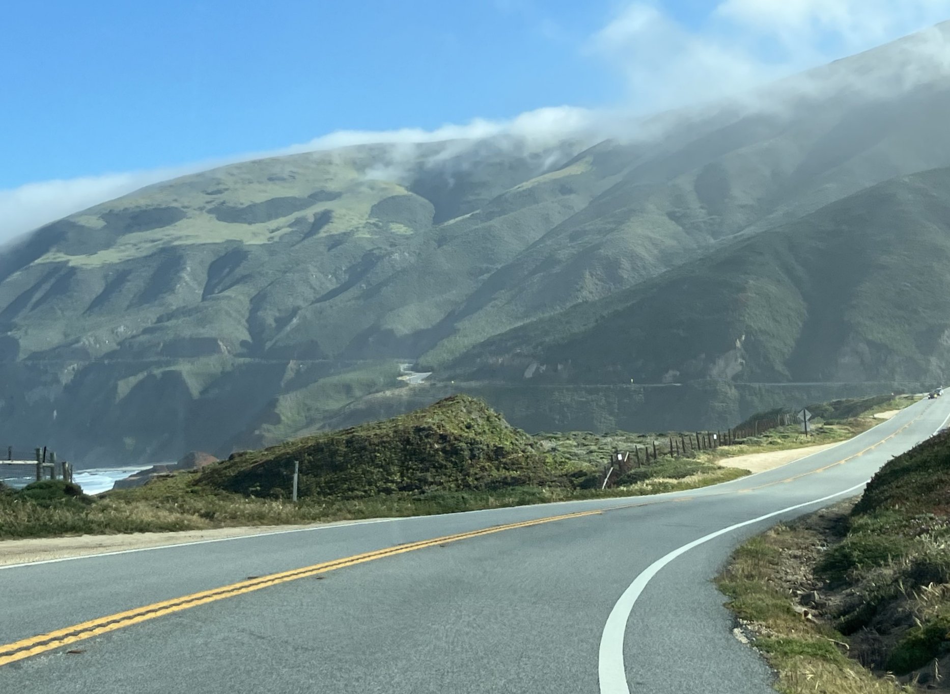 Mutkitteleva rannikkotie kaartuu vihreitä kukkuloita pitkin sinisen taivaan alla, ja matalat pilvet peittävät osittain vuorenrinteitä taustalla.