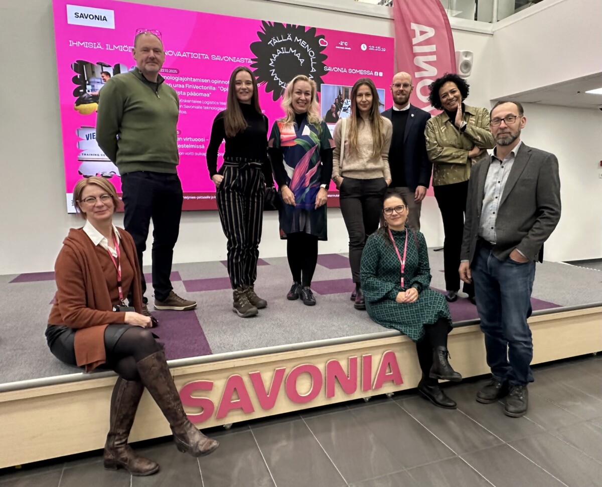 Yhdeksän ihmistä poseeraa ja hymyilee lavalla, jonka etupuolelle on kirjoitettu SAVONIA. Heidän takanaan näkyy kirkkaan vaaleanpunainen näyttö, jossa on tekstiä ja kuvia. Jotkut ihmiset seisovat, toiset istuvat tai kyykistyvät.