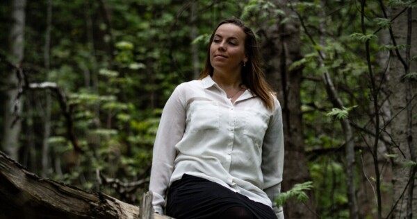 Valkoiseen puseroon ja mustaan hameeseen pukeutunut nainen istuu kaatuneen puun päällä tiheässä, vihreässä metsässä ja katselee sivulle rento ilme kasvoillaan. Tausta on täynnä puita ja lehtiä.