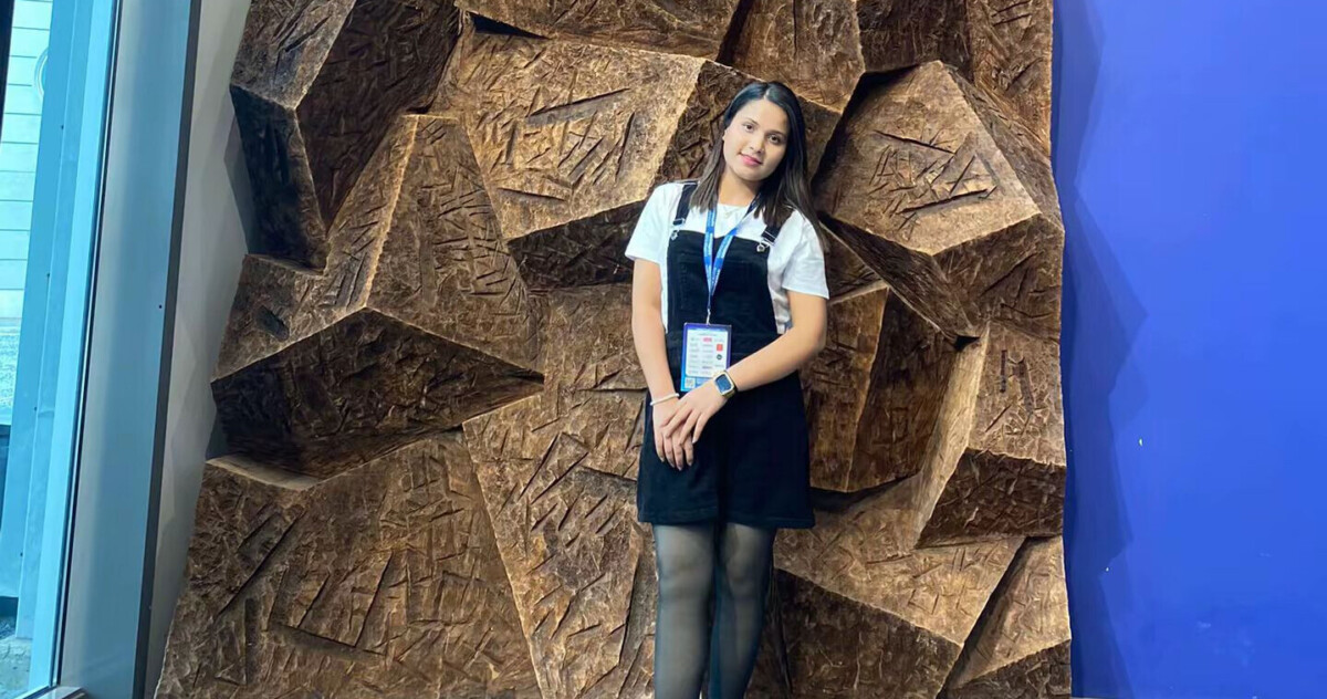 Mustaan mekkoon ja valkoiseen paitaan pukeutunut nainen poseeraa seinän edessä, jossa on suuria, kuvioituja, ruskeita geometrisia muotoja. Hänellä on yllään henkilökortilla varustettu kaulanauha ja hän seisoo kirkkaan sinisen seinän vieressä.