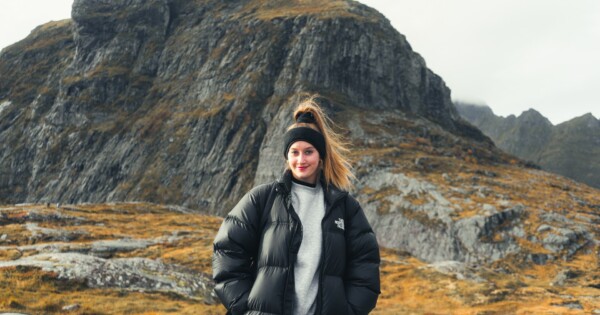 Mustaan puserotakkiin ja otsapantaan pukeutunut nainen seisoo hymyillen ulkona, taustalla kallioinen, ruohoinen vuori pilvisen taivaan alla.