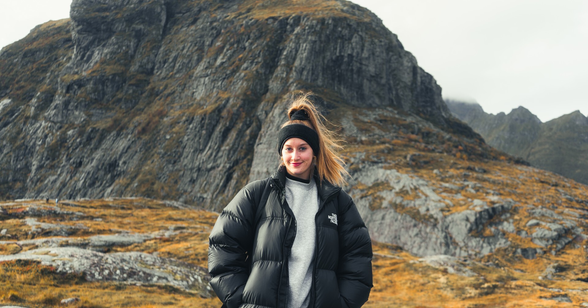 Mustaan puserotakkiin ja otsapantaan pukeutunut nainen seisoo hymyillen ulkona, taustalla kallioinen, ruohoinen vuori pilvisen taivaan alla.