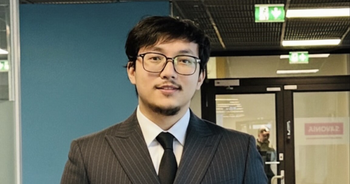 Silmälasipäinen mies, jolla on lyhyt parta, tumma neulenauhapuku ja musta solmio, seisoo sisätiloissa modernissa toimistossa, jonka taustalla on siniset seinät ja lasiovet.