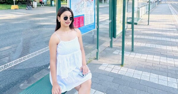 Valkoiseen mekkoon ja aurinkolaseihin pukeutunut nainen istuu vihreällä penkillä aurinkoisella bussipysäkillä, ja hänen takanaan on kaupungin kartta ja kadun toisella puolella näkyy puita.