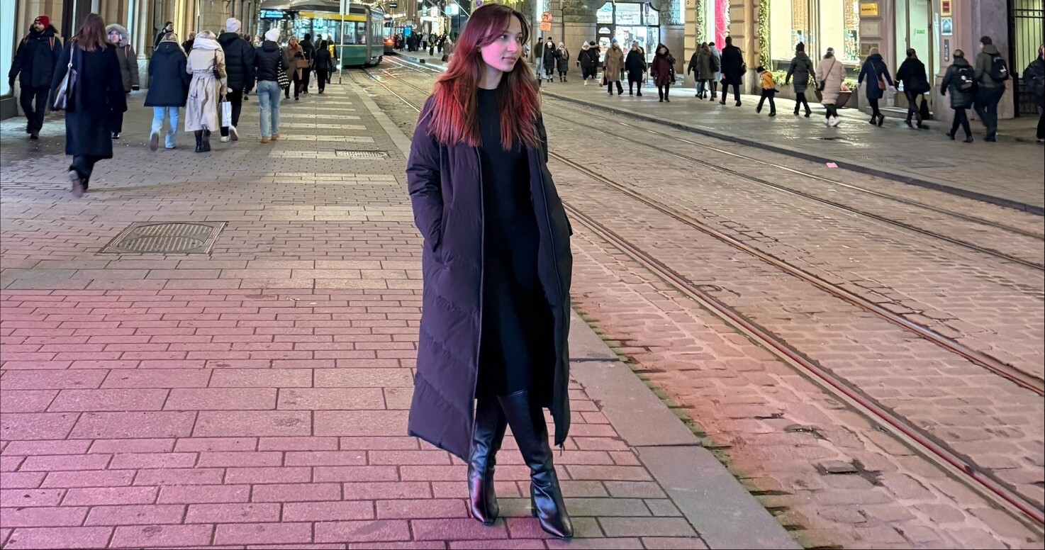 Mustaan pitkään takkiin ja saappaisiin pukeutunut nainen seisoo yöllisellä kaupunkikadulla, jonka taustalla näkyy raitiovaunukiskoja ja kirkkaissa valoissa käveleviä ihmisiä.