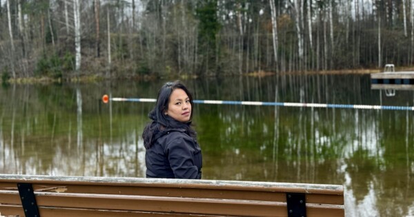 Tummiin takkeihin pukeutunut nainen istuu puisella penkillä rauhallisen järven rannalla ja kääntyy katsomaan kameraan. Taustalla on puita, joilla on paljaat oksat, ja veden päällä kelluva este.