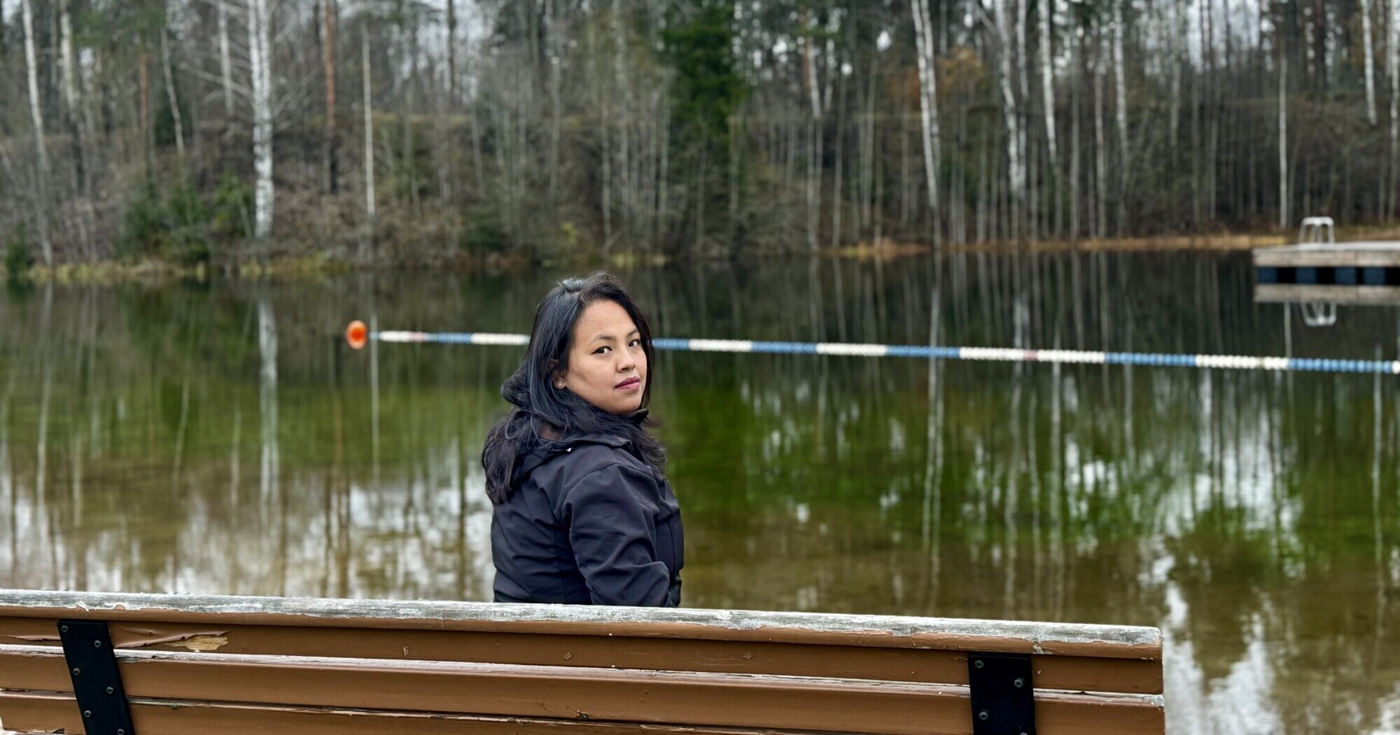 Tummiin takkeihin pukeutunut nainen istuu puisella penkillä rauhallisen järven rannalla ja kääntyy katsomaan kameraan. Taustalla on puita, joilla on paljaat oksat, ja veden päällä kelluva este.