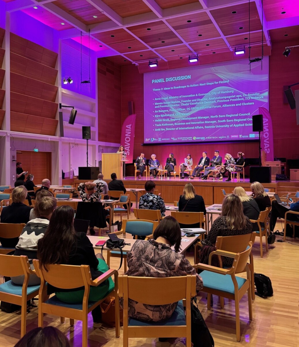 Paneelikeskustelu käydään lavalla modernissa auditoriossa, jossa on violetti valaistus. Yleisö istuu pöydissä paneeliin osallistuvia henkilöitä vastapäätä, jotka istuvat rivissä keskustelunaiheita näyttävän suuren näytön alla.