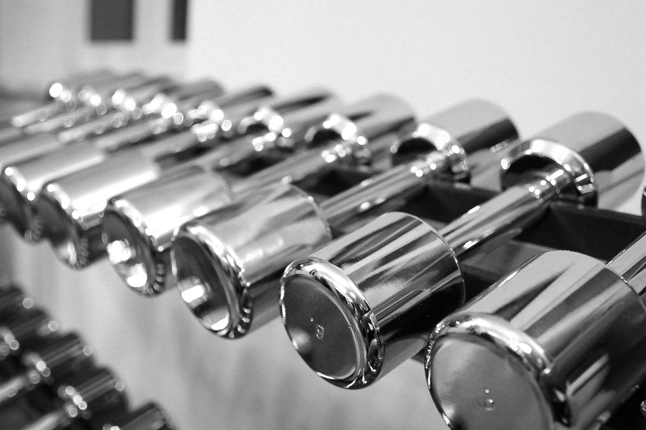 Rivi kiiltäviä, metallisia käsipainoja on järjestetty siististi kuntosalin telineeseen. Kuva on mustavalkoinen, mikä korostaa painojen heijastavia pintoja ja sylinterimäisiä muotoja.