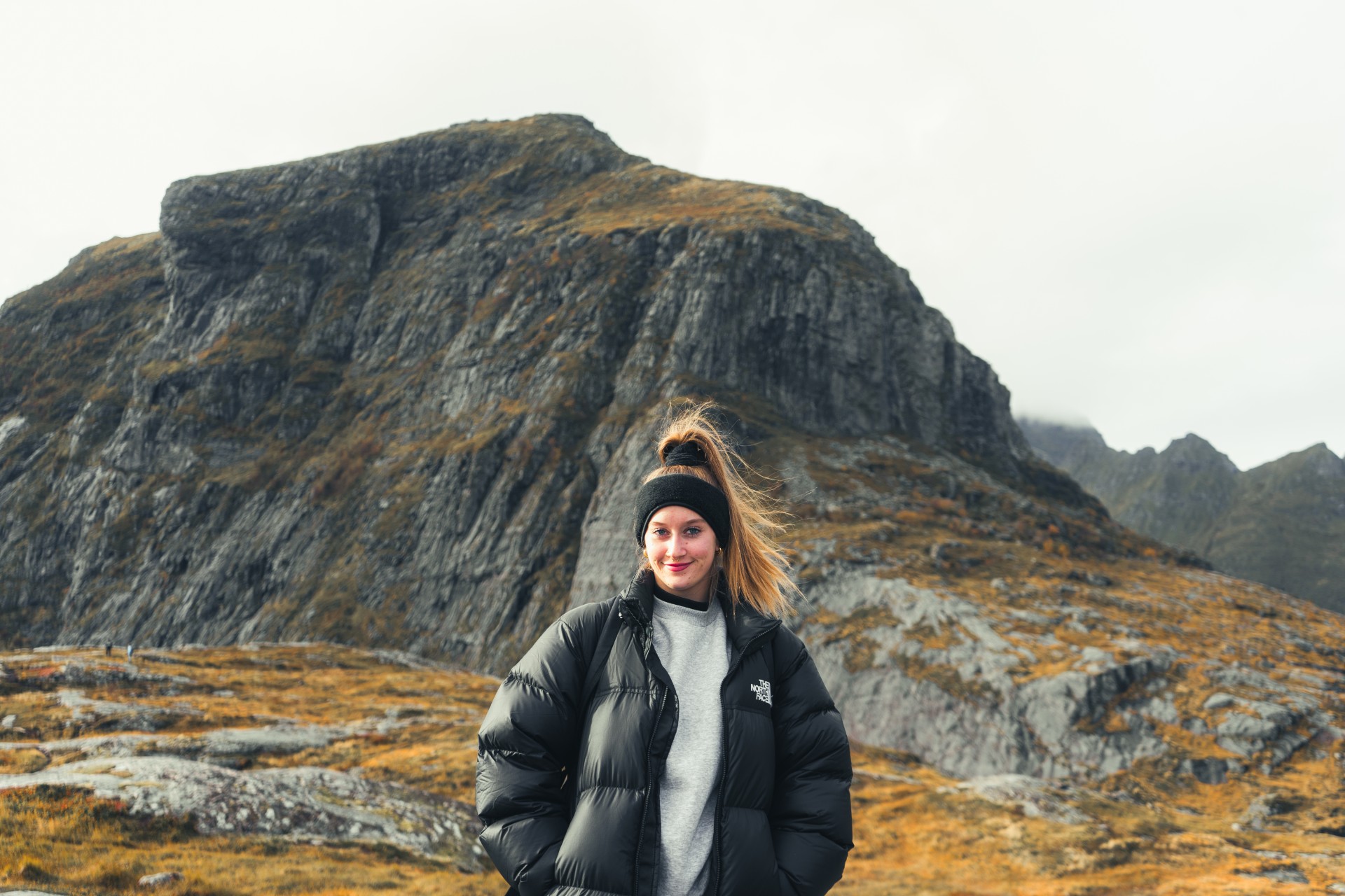 Mustaan puserotakkiin ja otsapantaan pukeutunut nainen seisoo hymyillen suuren kalliovuoren edessä, jonka alarinteillä on syksyistä ruohoa pilvisen taivaan alla.