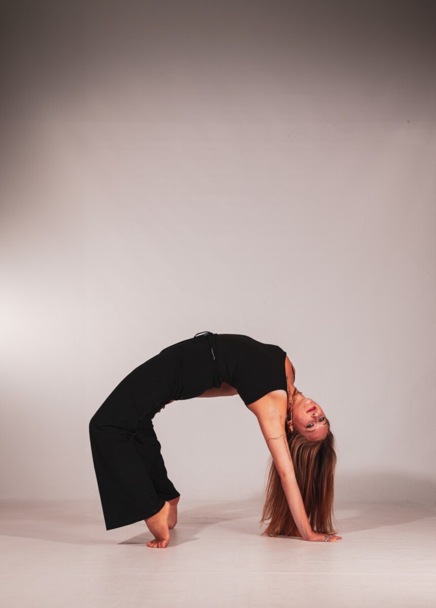 Mustiin vaatteisiin pukeutunut pitkähiuksinen nainen joogaa selinmakuulla, tasapainoilee käsillään ja jaloillaan pää taaksepäin kallistuneena minimalistisessa studiossa, jossa on neutraali tausta.