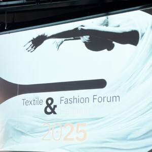 Valkokankaalla näytetään "Finnish Textile & Fashion: Textile & Fashion Forum Helsinki 2025" ja taiteellinen kuva henkilöstä, joka on verhoutunut virtaavaan kankaaseen. Luonnonvalo tulee sisään vasemmalla olevista ikkunoista.