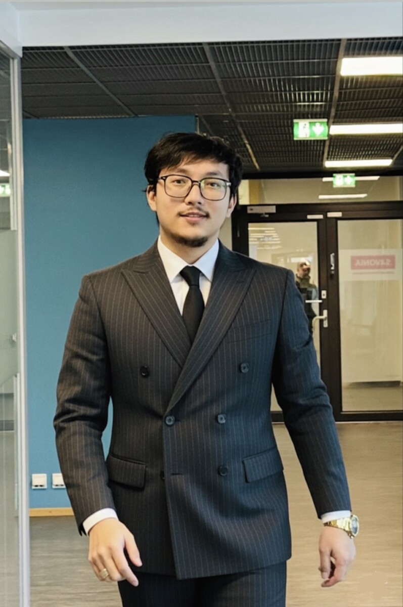 Tummahiuksinen ja silmälasipäinen mies, jolla on tumma neulenauhapuku, musta solmio ja kultakello, seisoo sisätiloissa modernissa toimistokäytävässä, jossa on siniset seinät ja lasiovet.